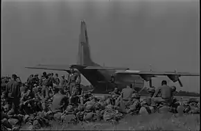 Troops from the 101st Airborne Division are pictured sitting in the grass at the Oxford airfield as a large military cargo aircraft unloads a Jeep