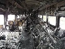 A rail passenger car interior with no upholstery, showing many burn marks and other fire damage. Several long pieces of metal are piled in the center near the back end of the car.