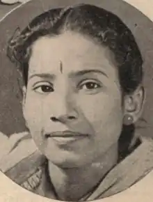 A young South Asian woman, dark hair parted center and dressed back behind her ears; she is wearing a light-colored shawl