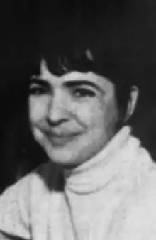 A smiling white woman with dark hair cut in a fringe, and dark eyes; wearing a light-colored turtleneck sweater