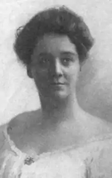 A white woman with dark hair in a bouffant updo, wearing a white blouse or dress with an off-the-shoulder neckline