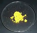 Some canary-yellow powder sits, mostly in lumps, on a laboratory watch glass.