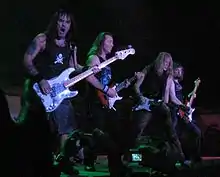 Four members of Iron Maiden are shown in concert. From left to right are a bass guitarist and then three electric guitarists. All members shown have long hair.