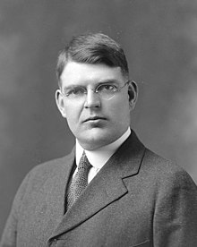 Posed photograph of a gentleman, standing, wearing a dark suit and glasses