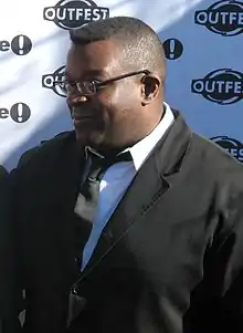 Isaac Julien facing to the left while wearing a white shirt, a black coat, and a black tie