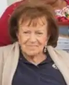 An older woman with olive skin and dark hair in bangs; she is wearing a dark shirt and a tan cardigan