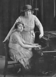 A young woman with dark bobbed hair is seated at a piano; an older woman wearing a brimmed hat is standing behind, one hand on chair, one hand on piano; both are wearing light-colored lacy gowns