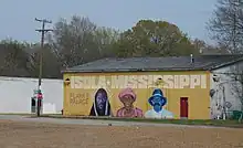 A mural on the side of a brick building, showing "ISOLA MISSISSIPPI", the headshots of three people, and "PLAYAS PALACE".