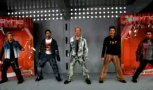 Five NSYNC members wearing prosthetic makeup resembling dolls, while standing in front of an enlarged orange cardboard package inside a silver shelf