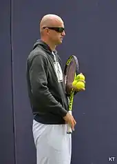 Ivan Ljubičić standing in athletic gear and sunglasses, holding a racquet in his right hand and three balls in his left hand.