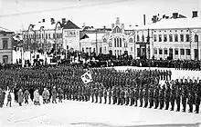 A parade of Finnish Jägers at the Vaasa town square. Spectators are gathered around the soldiers in the background. General Mannerheim is inspecting the formation in the foreground.