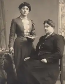 An image of two women. One is standing with a book in her left hand and is resting her right hand on a table. The other woman is seated in a wooden chair.