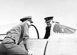 Two men in military uniforms with peaked caps on opposite sides of a military aircraft cockpit with open canopy