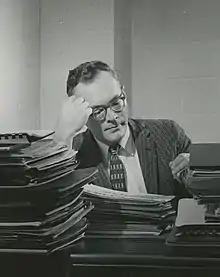 A man with glasses sitting down at a desk covered in papers and books