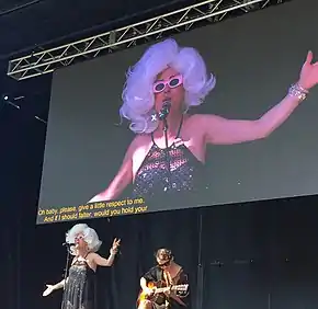 Bond, wearing a large white wig sings with Carerra, a guitar player, below a projected image of Bond