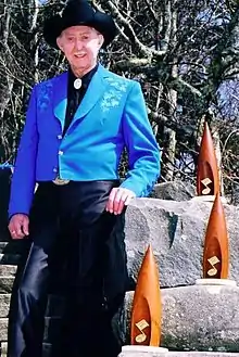 An old man in a black cowboy hat, blue jacket and black pants, standing next to some rocks on which a number of trophies are displayed
