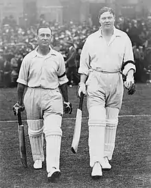 Two batsmen walk on to a cricket ground with a crowd behind them