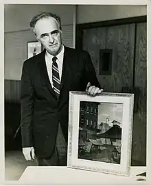 Black and white photograph of Jacob Glushakow, Chairman of JCC Art Committee. He is holding a painting on a table. The painting is a city scene, with small market stalls in front of a larger building.