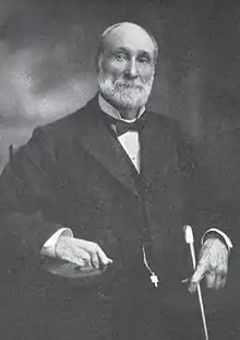 A man with receding gray hair and a gray beard and mustache. He is wearing a black coat, white shirt, and black bow tie