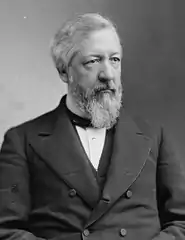 A bearded man with a solemn expression sits, while wearing a dark double breasted suit