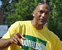 A man, wearing a yellow T-shirt, is standing and pointing to the camera.