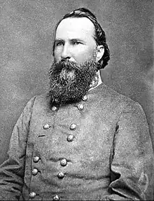Black and white photo shows a man with a thick beard and a receding hairline. He wears a gray military uniform with two rows of buttons.