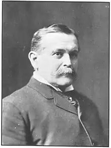 A bust-length black and white photo of a man in a tight collared jacket with short hair and a bushy mustache