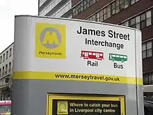 Large metal-skinned streetside sign painted silver, white and yellow indicating an interchange of rail and bus services