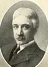 White male with white hair and a mustache in a dark suit