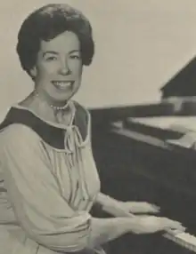 Black-and-white photograph of a brunette woman smiling at camera with her hands on the keyboard of a dark grand piano