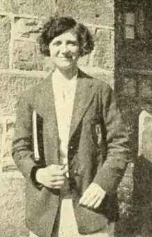 A smiling young white woman wearing a jacket over a white dress; photographed outdoors in front of a stone wall; she has a bobbed haircut and a book under one arm