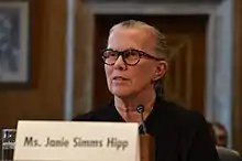 A woman with grey hair, tortoise shell colored glasses, and a black sweater sits behind a microphone and a name tag that reads "Ms. Janie Simms Hipp".