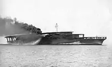 An aircraft carrier steaming at full speed. Thick black smoke billows from its curved smoke stack.