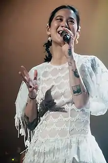 Zauner smiling while she performs in a white dress with a microphone in her left hand and several permanent tattoos on her arms