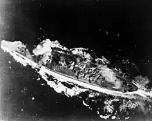 A close view of a large warship from almost directly overhead. Her wake is streaming out behind her and two trails of smoke are visible: a faint plume near her smokestack and a much thicker white plume partially obscuring her foremost main gun turret.