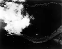 A distant overhead view of a patch of ocean; towards the left, partially obscured by a patch of cloud, is a large warship. The ship's wake trails out in a zig zag pattern behind her.