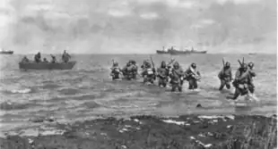 A black and white photograph of soldiers wading ashore from a small boat. Ships are visible in the distance.