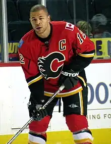 Hockey player in red and black uniform, with a "C" in the middle and no helmet on. He holds his stick diagonally.