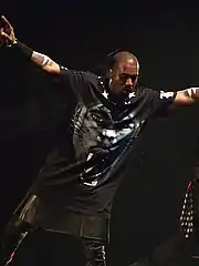 A man holding a microphone in his right hand whilst on stage. He wears a black T-shirt with a tiger-like face on it, tight leather pants, and a kilk.