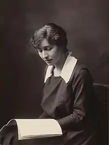 A white woman, seated, wearing a loose-fitting dark dress with a large white collar. She has an open soft-covered book or magazine in her lap, and is looking down at it.