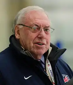 Elderly man with white hair and eyeglasses, wearing a navy blue jacket with a USA Hockey logo