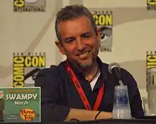 A Caucasian male sits at a desk, wearing a black shirt with a microphones in front of him.