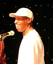 A dark-skinned man wearing a white baseball cap, singing into a microphone