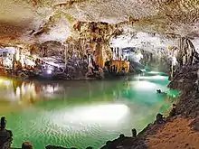 Brightly illuminated cave traversed by a bright emerald colored subterranean body of water encircling a variety of speleothems in the background