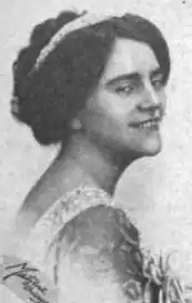 Photograph of a white woman, smiling over one shoulder; her dark hair is parted and dressed up with a tiara at the crown; she is wearing a gown with a low back.