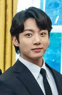 A close-up shot of Jungkook standing in a black-colored suit, looking directly at the camera.
