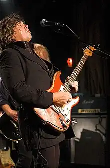 Dark haired man wearing dark clothing playing a right-handed electric guitar with another musician and amplifier behind