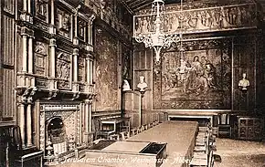 An elaborately-furnished room with a large fireplace, tapestry on the wall, and table and chairs in the centre.