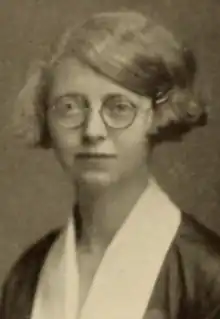 A young white woman with fair bobbed hair, wearing round glasses and a garment with white lapels