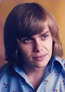 close-up of young white male with longish hair, wearing a patterned shirt, looking left of camera